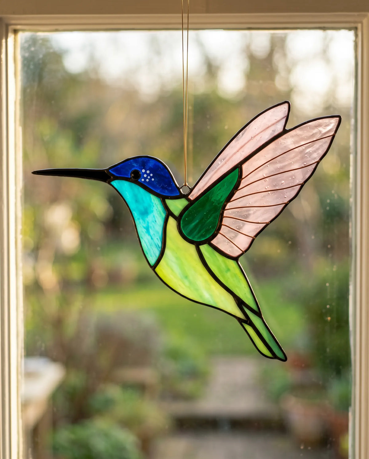 Iris's mouth-blown hummingbird suncatcher hanging in a sunlit Hertfordshire window, the rose-pink wing glowing translucent